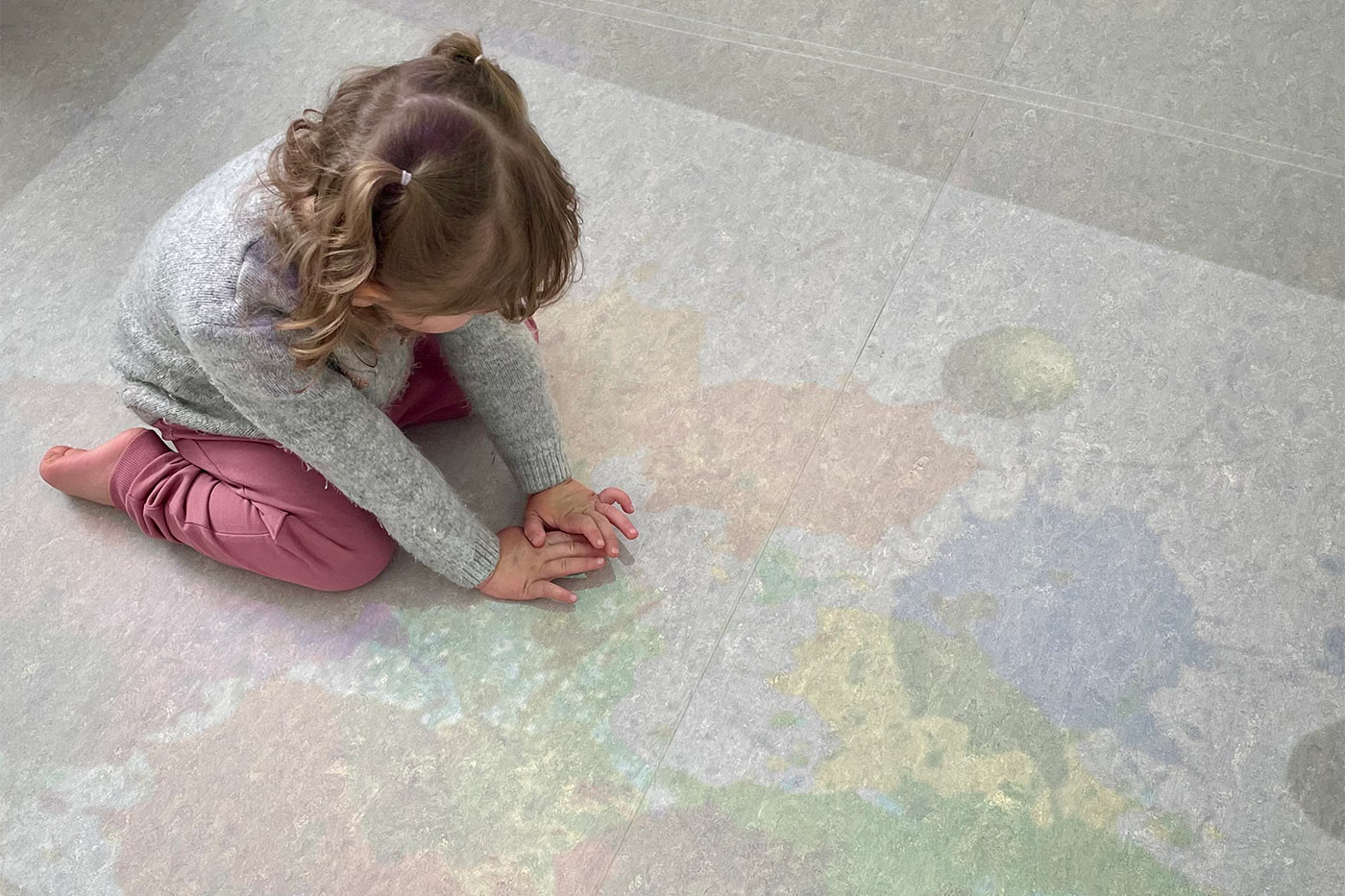 Kind kniet und berührt mit den Händen einen Farbklecks, der auf den Boden projiziert ist