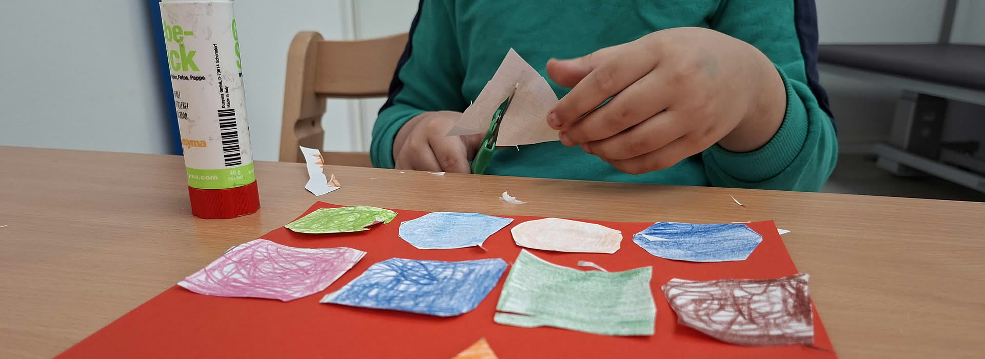 Bunte Papierformen in Kreis-, Dreieck- und Viereckform liegen auf rotem Papier, während ein Kind im Hintergrund mit einer Schere bastelt