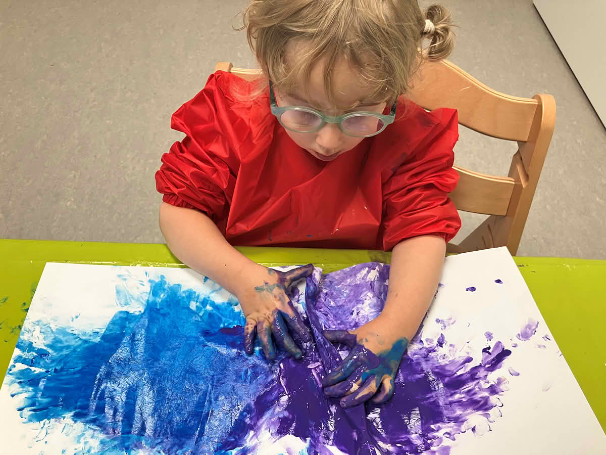 Kind gestaltet mit blauer und lila Fingerfarbe ein Blatt Papier